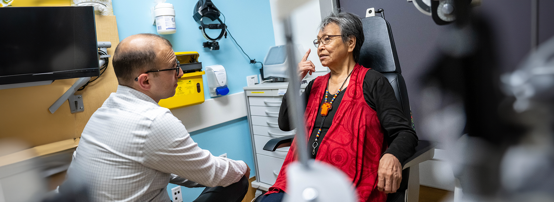 An older woman sits in an eye exam chair, speaking with a healthcare professional in a clinic. She is gesturing toward her eye while the provider listens attentively. Medical equipment and supplies are visible in the background.