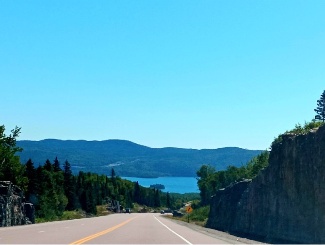 Photo of blue sky, moutains, blue water, green forests, and a road.