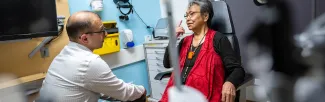 An older woman sits in an eye exam chair, speaking with a healthcare professional in a clinic. She is gesturing toward her eye while the provider listens attentively. Medical equipment and supplies are visible in the background.